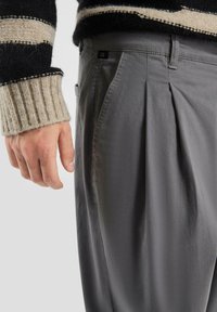 Pantalons plissés gris en tissu doux, avec une poche latérale et une étiquette noire. La texture est lisse et légèrement brillante.