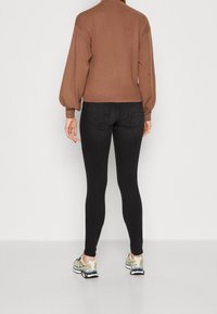 Brauner, langärmeliger Pullover mit enganliegendem Saum, kombiniert mit schwarzen Skinny Jeans und hellen Sportschuhen. Die Rückansicht zeigt voluminöse Ärmel.