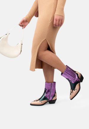 Ben i beige, ribbet midikjole med sideslids, iført lilla, sorte og beige ankelstøvler, holder en hvid håndtaske formet som en nyre.