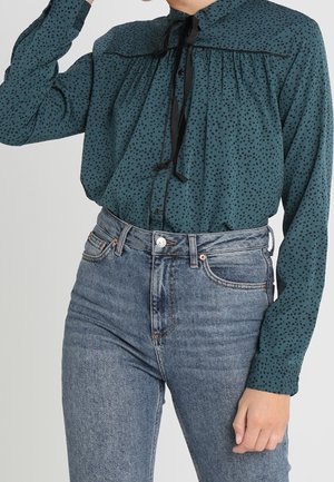 Kvinde iført en teal bluse med lange ærmer med sorte prikker og en bundet sløjfe ved kraven, kombineret med højtaljede blå jeans.