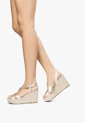 Zlaté kovové sandály na klínku s tkanou texturou platformy, které mají překřížené pásky a nastavitelnou přezku u kotníku. Otevřená špička.