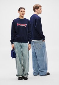 Tmavomodrá mikina s logom "tommy", v kombinácii s svetlomodrými voľnými džínsami. Modelka drží malú tmavomodrú kabelku a má na nohách čierne topánky.