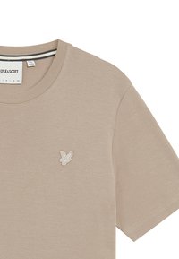 Beige Baumwoll-T-Shirt mit Rundhalsausschnitt und kurzen Ärmeln, das ein kleines, weiß gesticktes Logo auf der Brust trägt.
