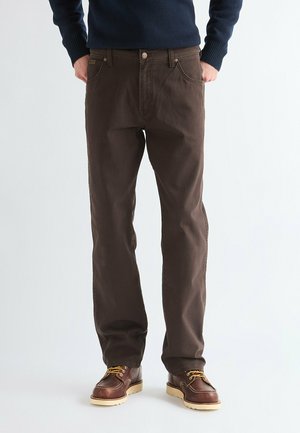 Man wearing dark brown pants and brown leather shoes with yellow laces, standing with hands in pockets against plain background.
