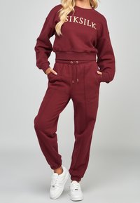 Conjunto de sudadera y pantalones de chándal en burdeos, fabricado en tela suave. La sudadera tiene un cuello redondo y un logo dorado. Los pantalones de chándal presentan una cintura elástica y puños.