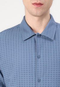Light blue button-up shirt featuring a textured fabric with a grid pattern, a structured collar, and two front buttons.