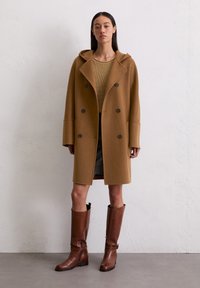 Manteau en laine marron avec capuche, design double boutonnage, grands boutons et manches larges, associé à des bottes en cuir marron montantes.