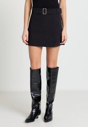 Donna con una minigonna nera con cintura e stivali neri in pelle testurizzata al ginocchio in piedi contro uno sfondo semplice.