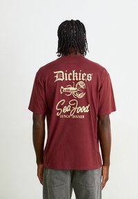 T-shirt en coton maroon avec un grand graphique de homard beige et le texte "Dickies SeaGood Lunch/Dinner" au dos. Manches courtes, coupe décontractée.