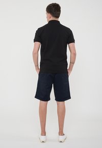 Polo shirt nero a maniche corte, abbinato a pantaloni corti blu navy. Modello visto di spalle, che indossa scarpe bianche. Design semplice e casual.