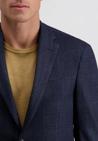 Mann, der ein strukturiertes, marineblaues Sakko mit einem subtilen Karomuster und einem senfgelben Strickoberteil trägt, von Hals bis zur Mitte des Oberkörpers zu sehen.