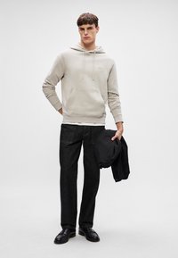 Beige huppari, jossa on etutasku ja kiristettävä huppu, yhdistetty tummansinisiiin farkkuihin ja mustiin kenkiin, takki kädessä.