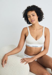 Φωτεινό μπεζ μπράλετ με λεία υφή, φαρδιές τιράντες και φαρδύ ιμάντα με λογότυπο. Συνδυασμένο με αντίστοιχα σλιπ, σε ένα απαλό, υφασμάτινο φόντο.