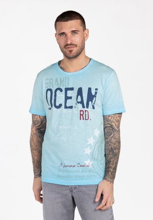 Mężczyzna z krótkimi włosami i tatuażami na rękawach ubrany w jasnoniebieską koszulkę z nadrukiem "Grand Ocean RD." oraz szare dżinsy, stoi na białym tle.