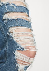 Blå slitna denimjeans med fransade knäfickor och flera revor. Texturen är grov, med lösa trådar och en bleknad look.