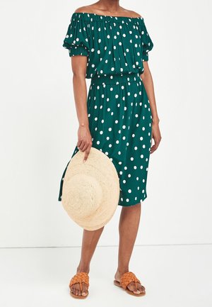 Woman wearing a green off-shoulder dress with white polka dots, holding a large woven sun hat, and wearing tan braided sandals.
