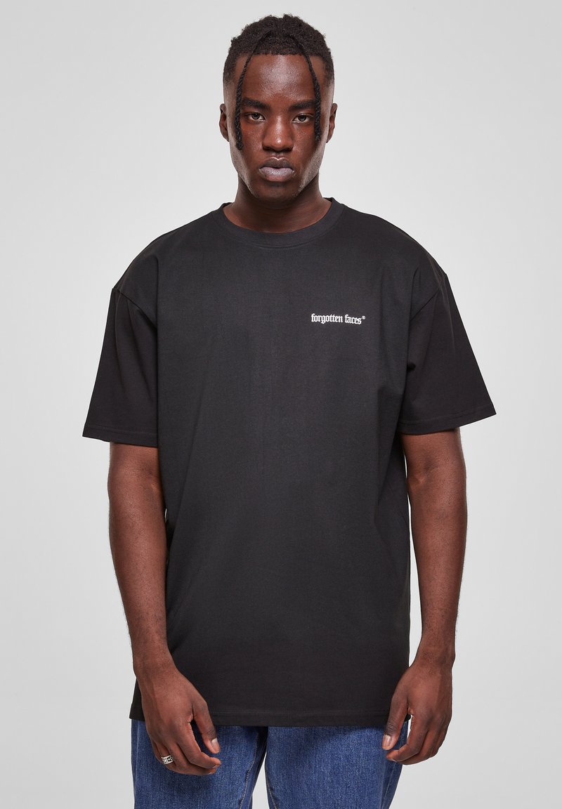 Svart bomull t-skjorte med korte ermer, løs passform, med "forgotten faces®" logo i hvitt på venstre bryst. Båret med blå jeans.
