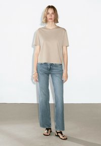 Beige T-Shirt mit kurzen Ärmeln, lockere Passform, kombiniert mit hellblauen Schlagjeans und schwarzen Sandalen mit dekorativem Hardware. Einfaches Design.