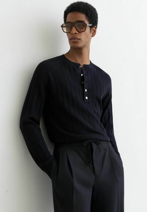 Mężczyzna w ciemnej prążkowanej bluzce z długim rękawem i guzikami oraz czarnych spodniach ze sznurkiem, stojący na tle białej ściany, ręce w kieszeniach.