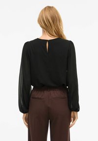 Blusa nera a maniche lunghe con maniche a sbuffo trasparenti, dettaglio sulla schiena a forma di chiave, abbinata a pantaloni bordeaux a vita alta con tasche laterali.