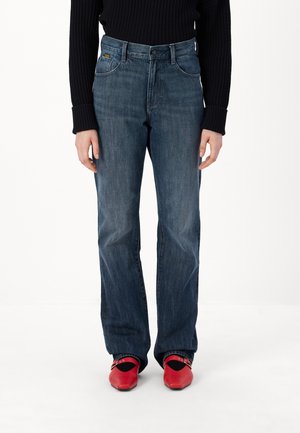 Osoba w ciemnoniebieskich dżinsach o prostych nogawkach, czarnym prążkowanym swetrze oraz jaskrawo czerwonych butach w szpic z czarnymi akcentami.