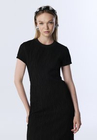 Zwart kort mouwen jurk met een gestructureerd, golvend patroon en een ronde halslijn. Accessoires omvatten een zwarte haarband en opvallende oorbellen.