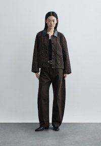 Brown patterned jacket with a black collar, large front pockets, paired with dark brown wide-leg pants and black shoes, standing on a gray floor.