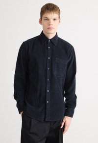 Camisa de pana azul marino con cuello abotonado, bolsillo frontal y botones azules; cuenta con un tejido texturizado y un ajuste entallado.