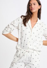 Weißes Schlafanzug-Set mit einer knopfbaren Bluse und elastischen Hosen, verziert mit kleinen, mehrfarbigen Herzmustern. Weicher Stoff.