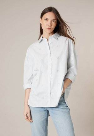 Femme aux longs cheveux foncés portant une grande chemise blanche à rayures bleues avec manches retroussées et un jean bleu clair, debout devant un fond beige.