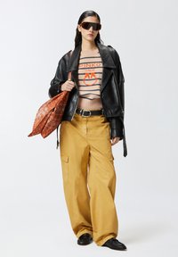 Crna kožna bikerska jakna, prugasti bež džemper s crvenim logom, žute široke hlače, smeša torba s uzorkom, tamne sunčane naočale, crne cipele.