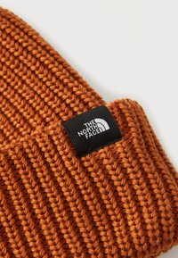 Orange knitted beanie with a ribbed texture. Features a black label with white text displaying "THE NORTH FACE."