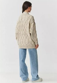 Beżowy sweter w warkoczowy splot o luźnym kroju, z teksturowanym wzorem. Połączony z jasnoniebieskimi spodniami o szerokich nogawkach i białymi trampkami.