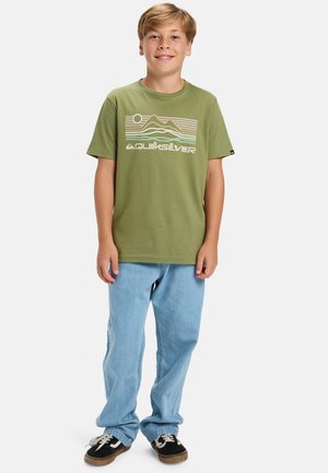 Olivově zelené tričko s bílým grafickým designem zobrazujícím hory a textem "Quiksilver". Nosené s světle modrými džíny a černými teniskami.