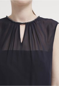 Blusa blu navy con scollo arricciato e apertura a chiave, caratterizzata da una rifinitura decorata con perline. Realizzata in un tessuto leggero e trasparente.