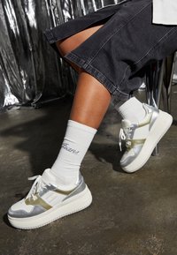 Białe i srebrne sneakersy z masywną podeszwą, ozdobione metalicznymi akcentami i gładką powierzchnią, noszone ze białymi skarpetami z napisem.