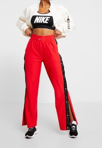 Pantalones deportivos rojos con aberturas laterales y ribete negro, con el logo Nike Pro, combinados con un crop top negro de Nike y una chaqueta bomber ligera.