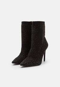 Botas de tobillo de ante negro con punta afilada, tacones de aguja y adornos con tachuelas que aportan una superficie texturizada. Cierre con cremallera en la parte trasera.