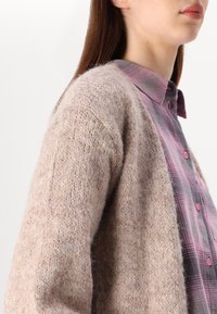 Kobieta w miękkim beżowym swetrze nałożonym na różowo-szarą koszulę w kratę zapinaną na guziki, widziana od ramienia do podbródka.