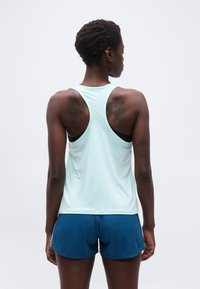 Femme aux cheveux courts, vue de dos, portant un débardeur bleu clair à dos nageur, un soutien-gorge de sport noir et un short de sport bleu.