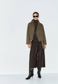Giacca in suede verde oliva con colletto alto, dolcevita nero, pantaloni a gamba larga marroni e stivali neri con tacco. Occhiali da sole scuri completano il look.