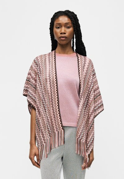 Femme aux longs cheveux tressés portant un châle rose à motif en zigzag par-dessus un haut rose clair et un pantalon gris, debout devant un fond uni.