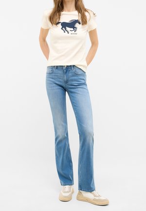 Kobieta ubrana w kremowy t-shirt z granatowym logo konia, jasnoniebieskie jeansy o prostych nogawkach oraz beżowo-białe trampki, stoi z rękami za plecami.