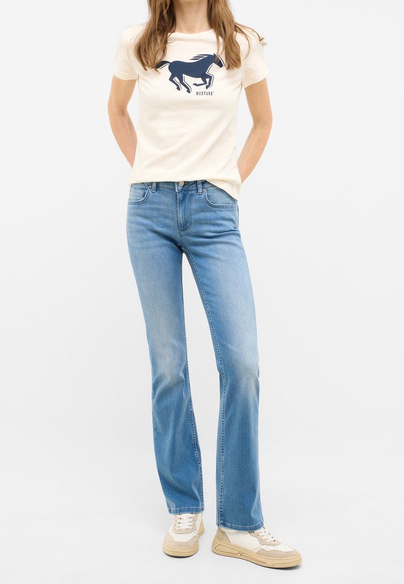 Femme portant un t-shirt crème avec un logo cheval bleu marine, un jean droit bleu clair et des baskets beige et blanches, debout avec les mains dans le dos.