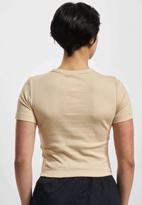 Person med kort svart hår som har på seg en tettsittende beige t-skjorte med korte ermer og svarte bukser, vendt bort fra en enkel hvit bakgrunn.