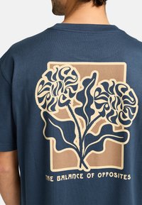 Marineblaues T-Shirt mit einem großen grafischen Motiv von zwei stilisierten Blumen mit geschwungenen Linien und Blättern, vor einem beigen rechteckigen Hintergrund.
