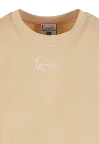 Vaalea beige puuvillaneule, jossa on ribbaus miehustassa ja valkoinen brodeerattu logo vasemmassa rinnassa. Pehmeä rakenne ja rento istuvuus.