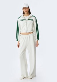 Witte zip-up jas met groene strepen en tekst, gecombineerd met losse witte broeken. De outfit heeft een zachte textuur en een casual pasvorm, aangevuld met een witte pet.