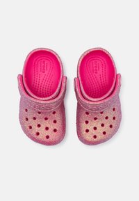 Zoccoli Crocs glitterati rosa con una finitura liscia e lucida; presentano fori di ventilazione e una fascia curva sul tallone per una calzata sicura e comfort.
