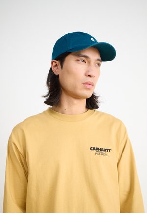 Türkise Kappe mit gebogenem Schirm und Logo; langärmliges senfgelbes Shirt mit schwarzem Text; glattes Gewebe, lockere Passform. Neutraler Hintergrund.